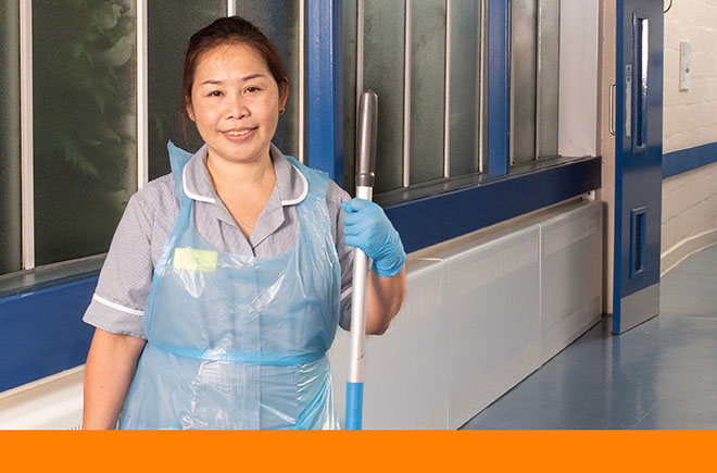 Smiling young woman in uniform and plastic apron stands in hospital corridor with mop